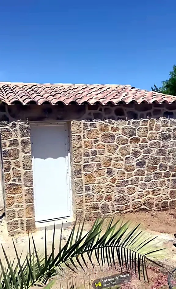 Cabanon aux murs façon pierre sèche, toit en tuiles, palmier au premier plan